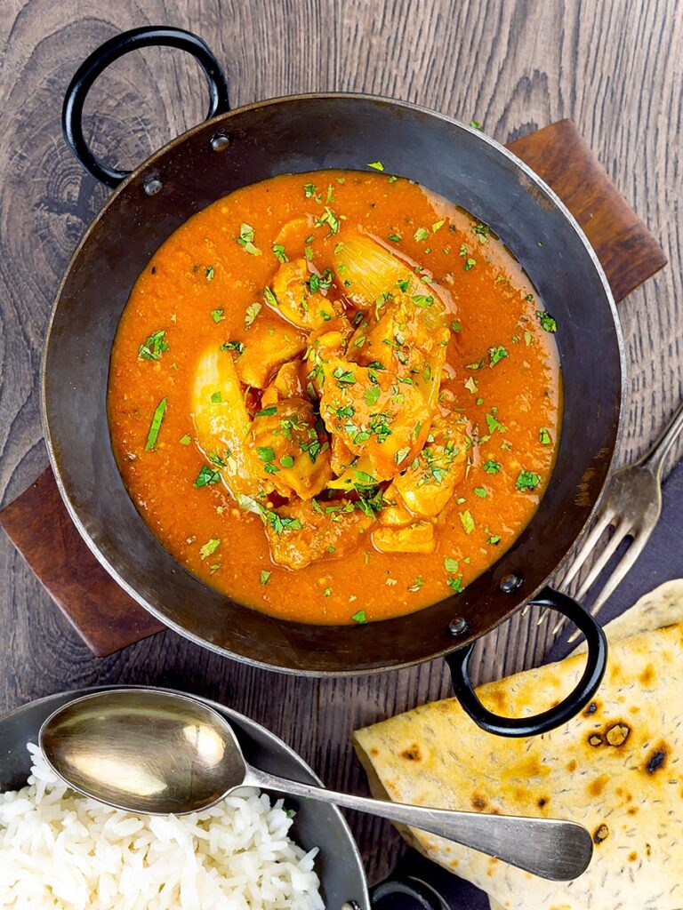 Portrait overhead image of a chicken jalfrezi curry with a coriander garnish served in an iron karahi bowl and chapatis and boiled rice