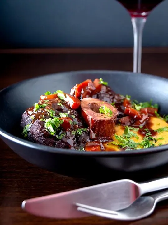 Portrait image of a beef shin osso bucco recipe served on polenta in a black bowl
