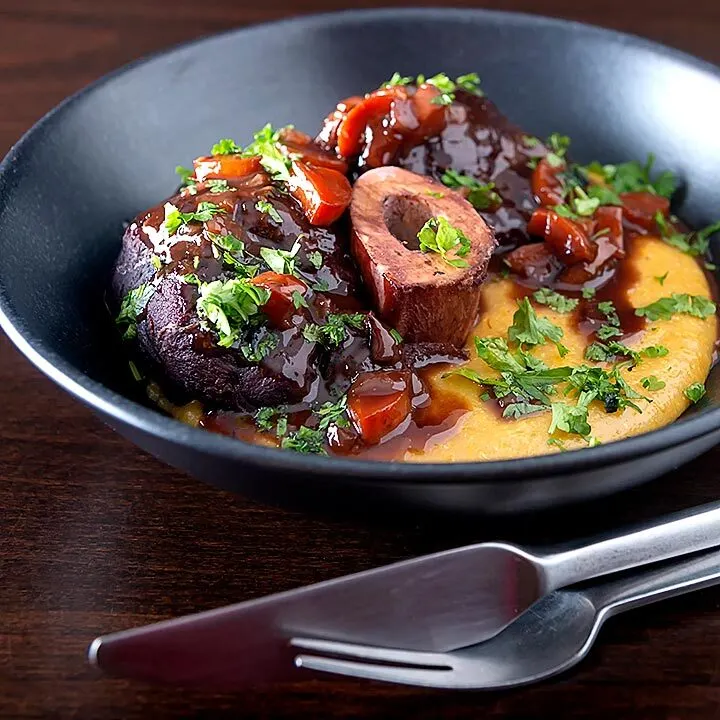 Square image of a beef shin osso bucco recipe served on polenta in a black bowl