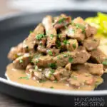 Portrait image of a Hungarian Pork Stew called Borsos Tokany served on a black plate with nokedli noodles and a text overlay