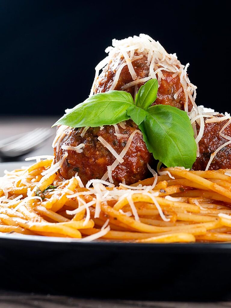 Portrait image of spaghetti and meatballs served with fresh basil and grated Parmesan cheese served on a white plate against a dark backdrop