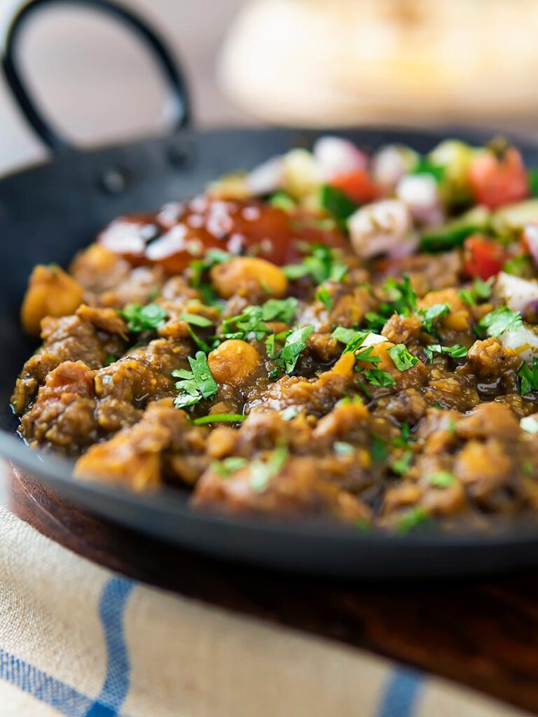 Portrait image of a vegan chickpea and lentil curry served in an iron kadai with mango chutney and kachumber salad