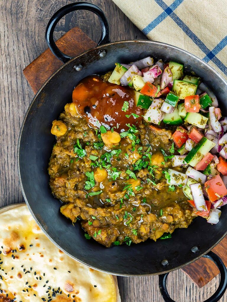 Portrait overhead image of a vegan chickpea and lentil curry served in an iron kadai with mango chutney and kachumber salad and chapatis