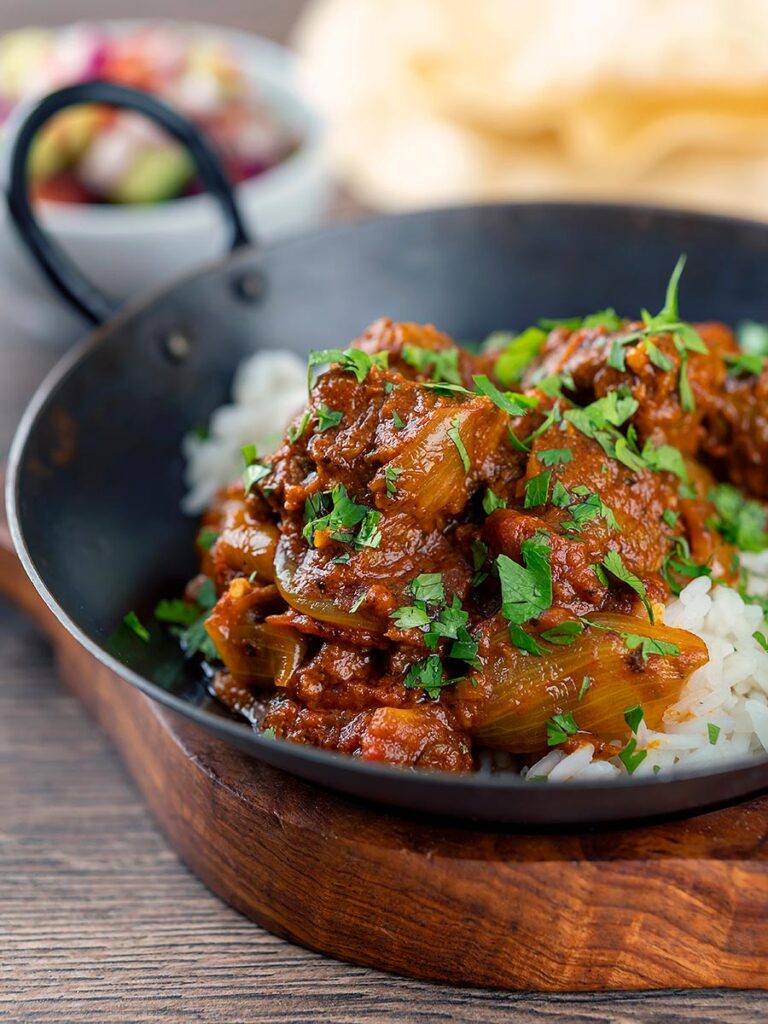 Portrait image of a beef bhuna curry served in an iron kadai with poppadoms and kachumber salad out of focus in the background