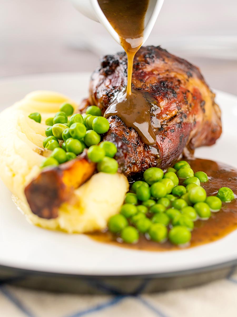 Portrait image of gravy being poured over oven braised minted lamb shanks served with mashed potatoes and peas