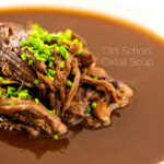 Portrait close up image of a British oxtail soup with a pile of shredded oxtail and snipped chives featuring a title overlay