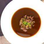 Portrait overhead image of a British oxtail soup with a pile of shredded oxtail and snipped chives