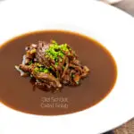 Portrait image of a British oxtail soup with a pile of shredded oxtail and snipped chives featuring a title overlay