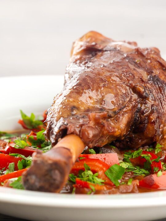 Portrait close up image of a slow cooker braised lamb shank served in a shallow white bowl with red peppers in a red wine gravy