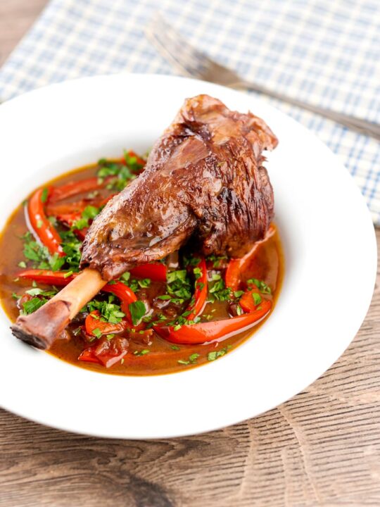 Portrait image of a slow cooker braised lamb shank served in a shallow white bowl with red peppers in a red wine gravy
