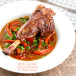 Portrait image of a slow cooker braised lamb shank served in a shallow white bowl with red peppers in a red wine gravy featuring a title overlay
