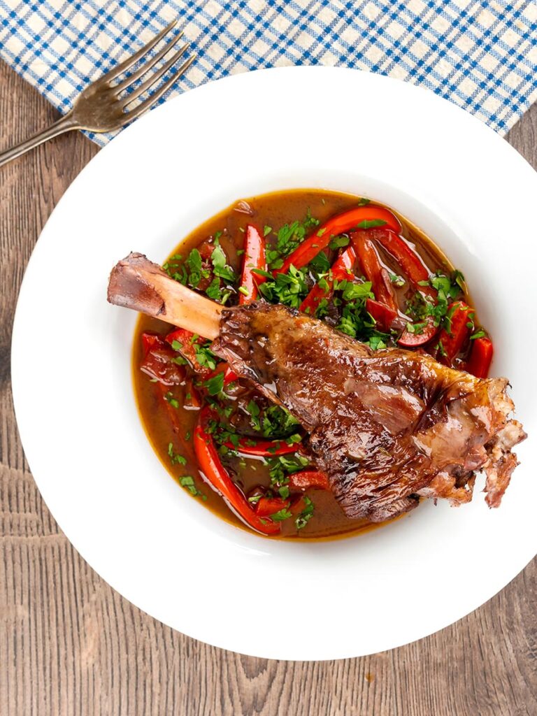 Portrait overhead image of a slow cooker braised lamb shank served in a shallow white bowl with red peppers in a red wine gravy