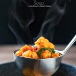 Portrait image of a steaming hot spicy vegetable soup with sweet potato, cauliflower and in a harissa broth in a ladle featuring a title overlay