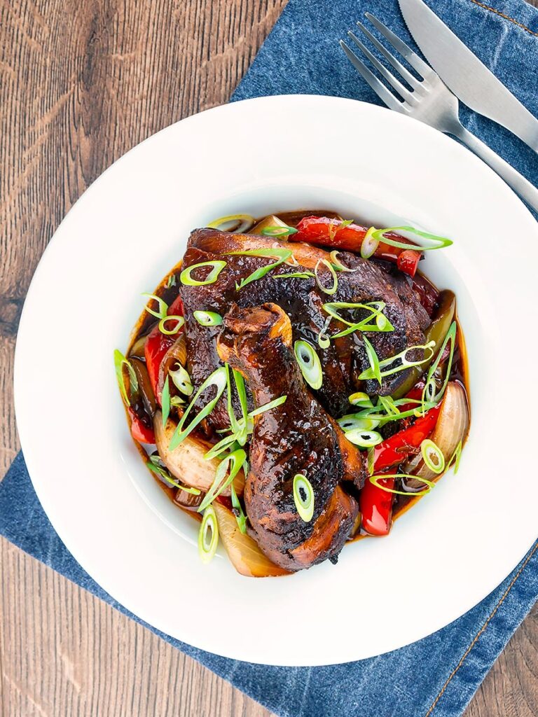 Portrait overhead image of a Jamaican brown stew chicken served in a white shallow bowl with a spring onion garnish