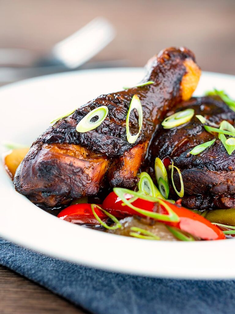 Portrait image of a Jamaican brown stew chicken served in a white shallow bowl with a spring onion garnish