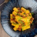 Portrait overhead image of a shredded duck ragu served with rigatoni pasta in a mottled dark blue pasta bowl