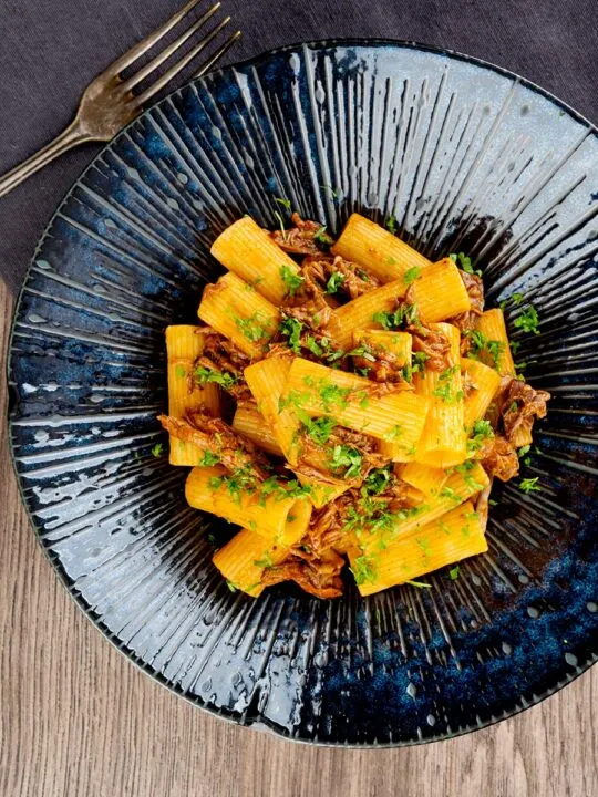 Portrait overhead image of a shredded duck ragu served with rigatoni pasta in a mottled dark blue pasta bowl