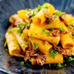Portrait image of a shredded Venetian duck ragu served with rigatoni pasta in a mottled dark blue pasta bowl