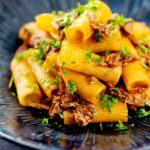 Portrait image of a shredded duck ragu served with rigatoni pasta in a mottled dark blue pasta bowl featuring a text overlay