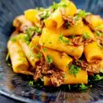 Portrait image of a shredded duck ragu served with rigatoni pasta in a mottled dark blue pasta bowl featuring a text overlay