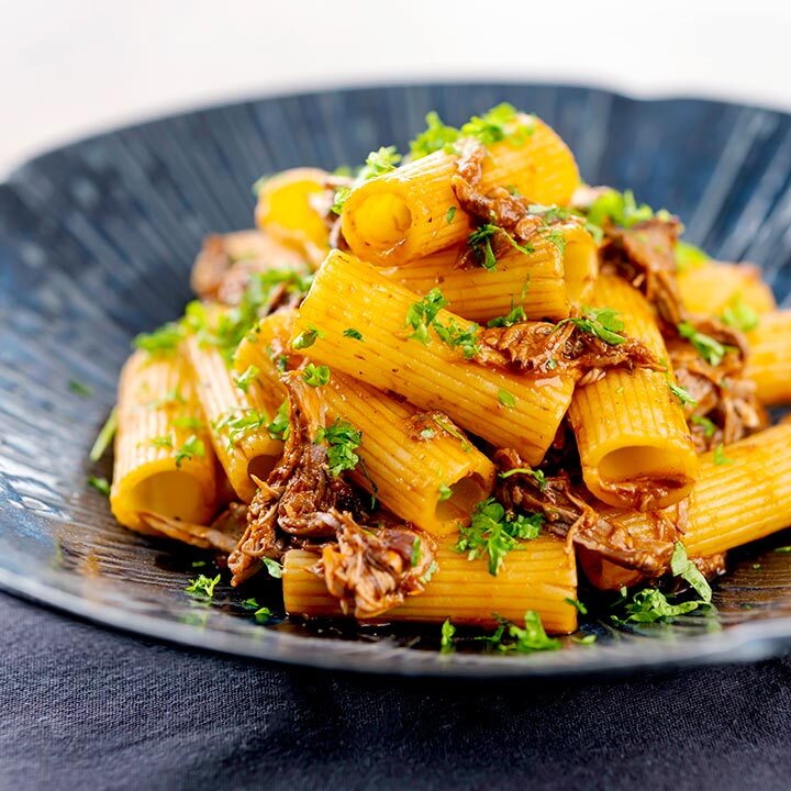 Square image of a shredded Venetian duck ragu served with rigatoni pasta in a mottled dark blue pasta bowl