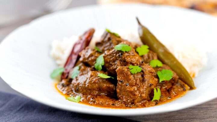 Portrait image of an achar gosht lamb or mutton curry served on a white plate with whole chilies, basmati rice and coriander leaves