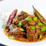 Portrait image of an achar gosht curry served on a white plate with whole chilies, basmati rice