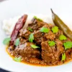 Portrait image of an achar gosht curry served on a white plate with whole chilies, basmati rice