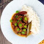 Portrait overhead image of an achar gosht curry served on a white plate with whole chilies, basmati rice and a naan bread