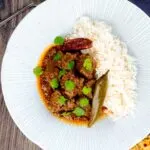 Portrait overhead image of an achar gosht curry served on a white plate with whole chilies, basmati rice and a naan bread