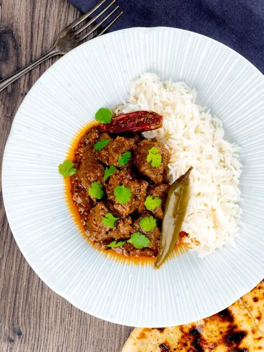 Portrait overhead image of an achar gosht curry served on a white plate with whole chilies, basmati rice and a naan bread