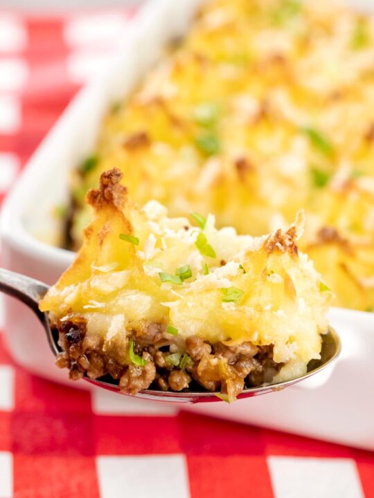 Portrait close up image of a spoonful of cheesy cumberland pie with a whole pie served in a white gratin dish in the background