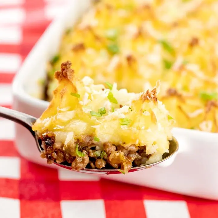 Square close up image of a spoonful of cheesy cumberland pie with a whole pie served in a white gratin dish in the background