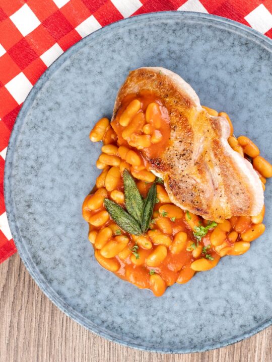 Portrait overhead image of Fagioli all’uccelletto or Italian Baked Beans served with a pork loin steak and crispy fried sage leaves