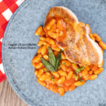 Portrait overhead image of Fagioli all’uccelletto or Italian Baked Beans served with a pork loin steak and crispy fried sage leaves featuring a title overlay