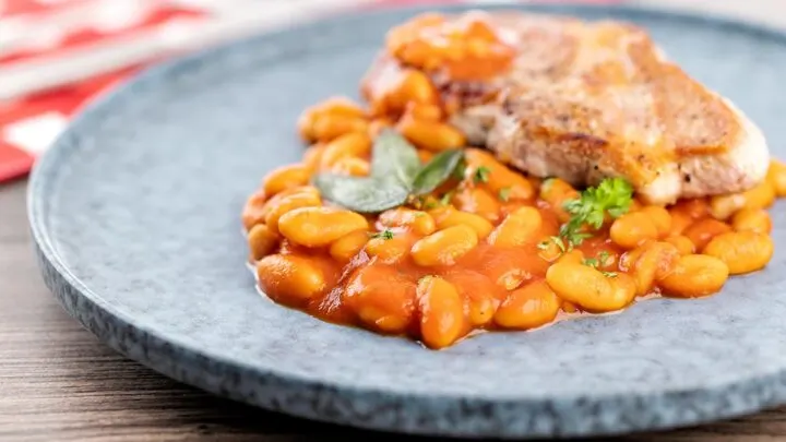 Landscape image of Fagioli all’uccelletto or Italian Baked Beans served with a pork loin steak and crispy fried sage leaves