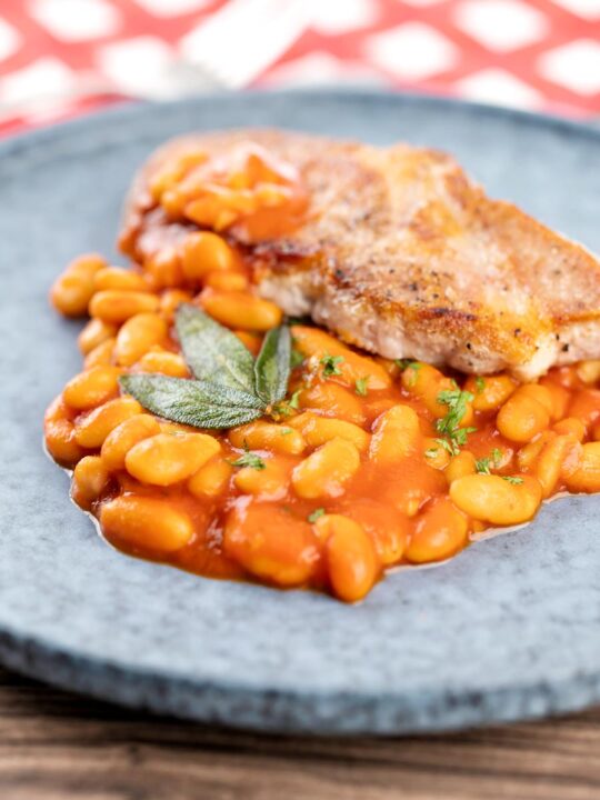 Portrait image of Fagioli all’uccelletto or Italian Baked Beans served with a pork loin steak and crispy fried sage leaves