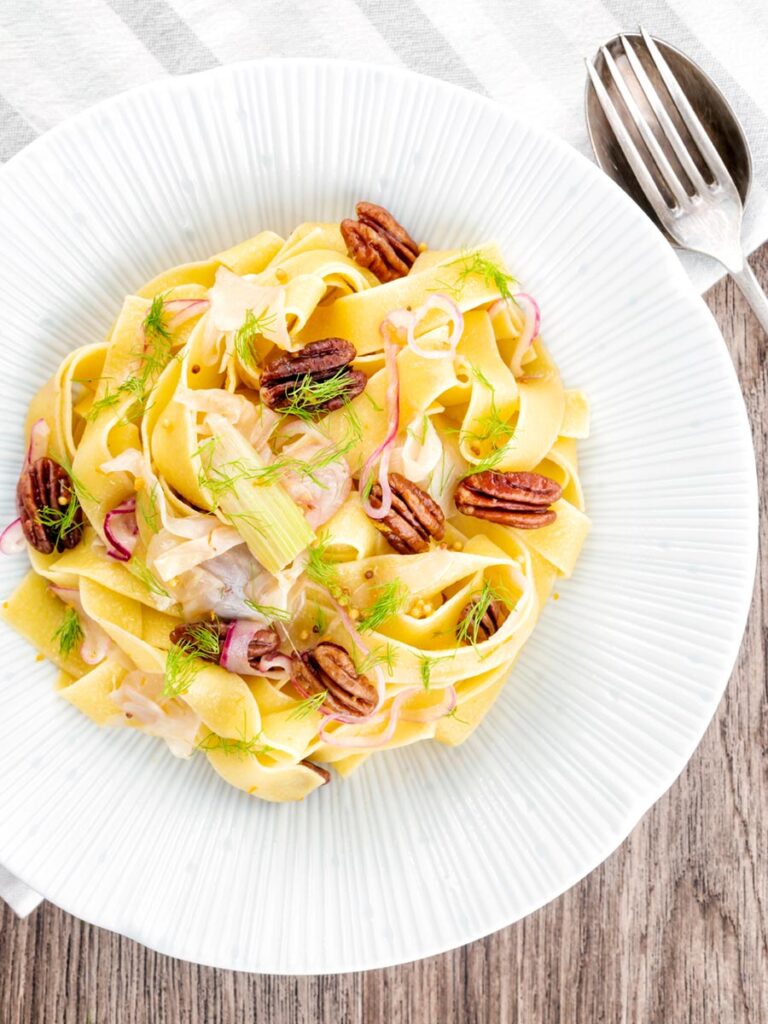 Portrait overhead image of fennel pasta made with pappardelle served with pecan nuts and red onion served in a white pasta bowl