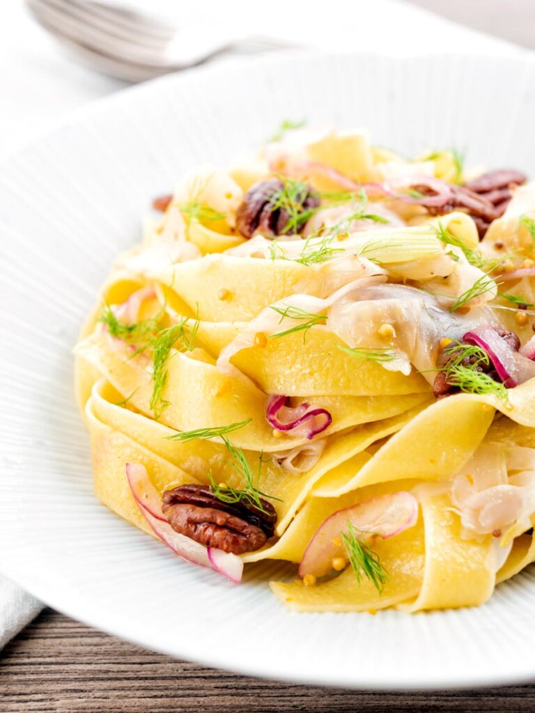 Portrait image of fennel pasta made with pappardelle served with pecan nuts and red onion served in a white pasta bowl