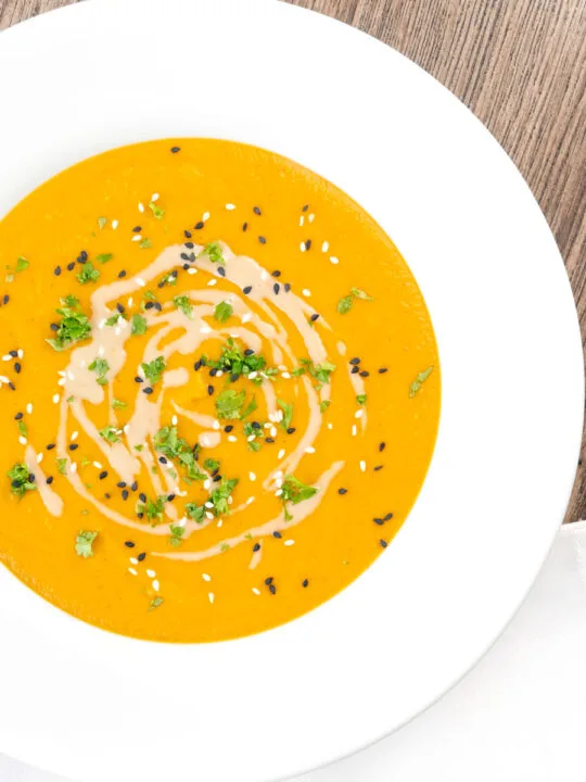 Portrait overhead image of a spicy carrot and lentil soup spiced with harissa paste and garnished with a swirl of tahini served in a white bowl