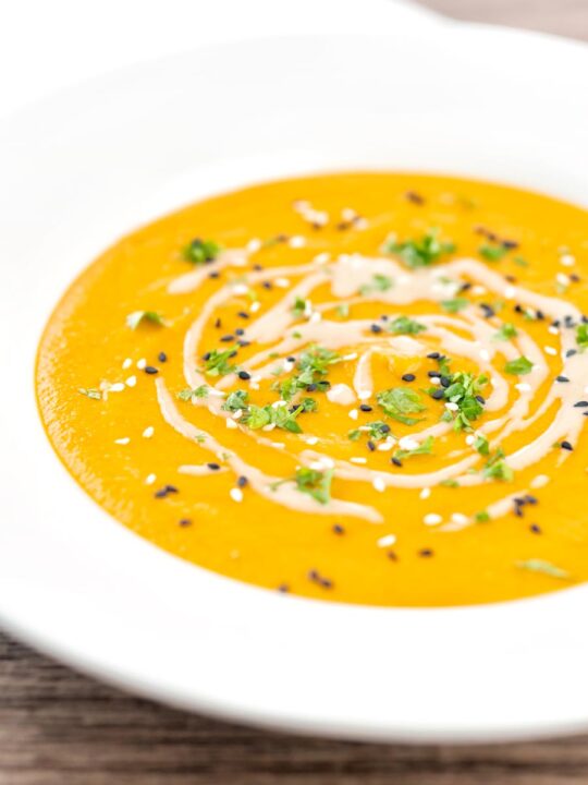 Portrait image of a spicy carrot and lentil soup spiced with harissa paste and garnished with a swirl of tahini served in a white bowl