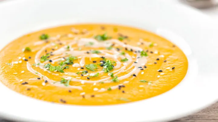 Landscape image of a spicy carrot and lentil soup spiced with harissa paste and garnished with a swirl of tahini served in a white bowl