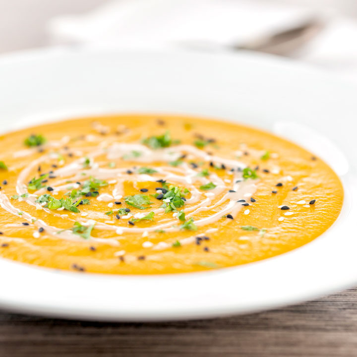 Square image of a spicy carrot and lentil soup spiced with harissa paste and garnished with a swirl of tahini served in a white bowl