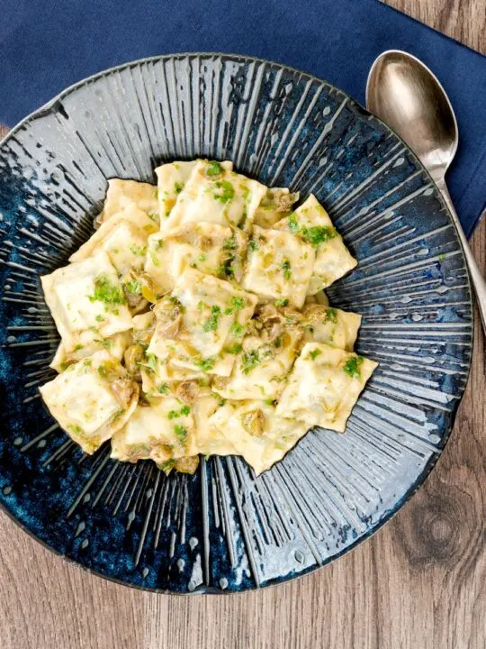 Portrait overhead image of homemade spinach and ricotta ravioli with a lemon caper butter sauce in a blue pasta bowl