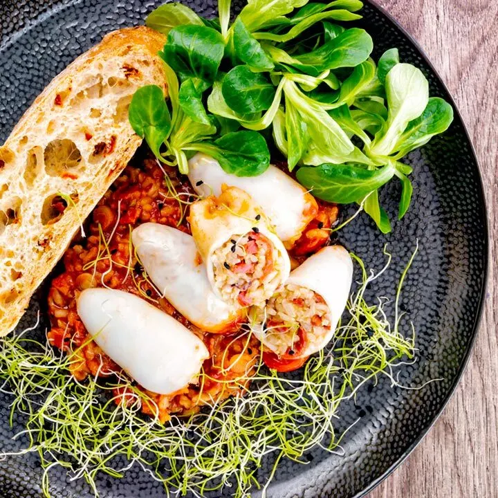 Square overhead image of brown rice stuffed squid on a tomato sauce served with ciabatta bread, lambs lettuce and sprouted onion seeds