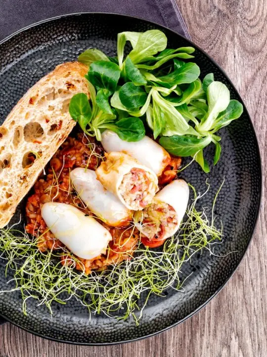 Square overhead image of brown rice stuffed squid on a tomato sauce served with ciabatta bread, lambs lettuce and sprouted onion seeds