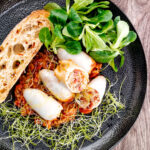 Portrait overhead image of brown rice stuffed squid on a tomato sauce served with ciabatta bread, lambs lettuce and sprouted onion seeds featuring a title overlay