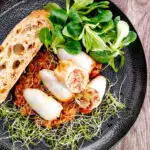 Portrait overhead image of brown rice stuffed squid on a tomato sauce served with ciabatta bread, lambs lettuce and sprouted onion seeds featuring a title overlay