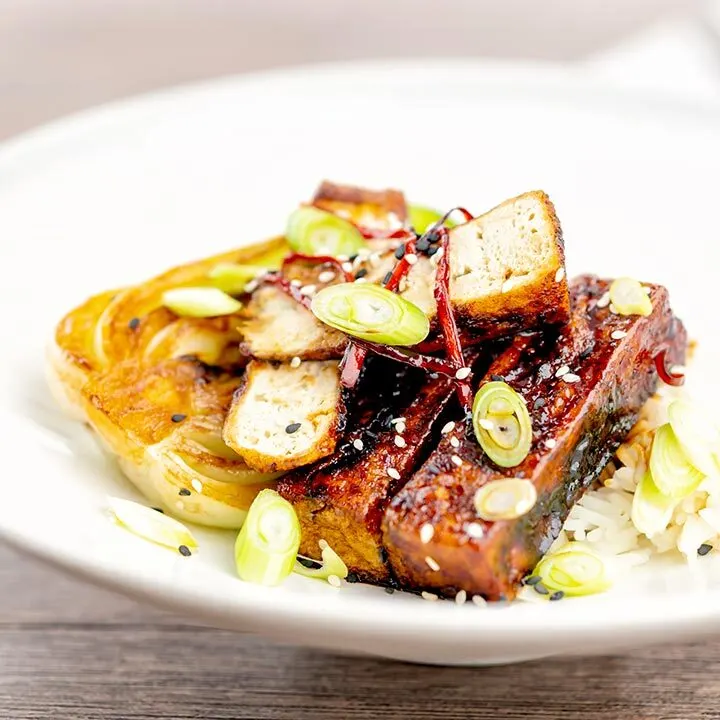 Square image of teriyaki tofu served with pak choi, white rice and spring onions on a white plate
