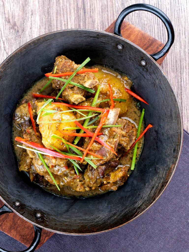 Portrait overhead image of Jamaican curry goat with potatoes served with shredded chilli and green onions served in an iron bowl